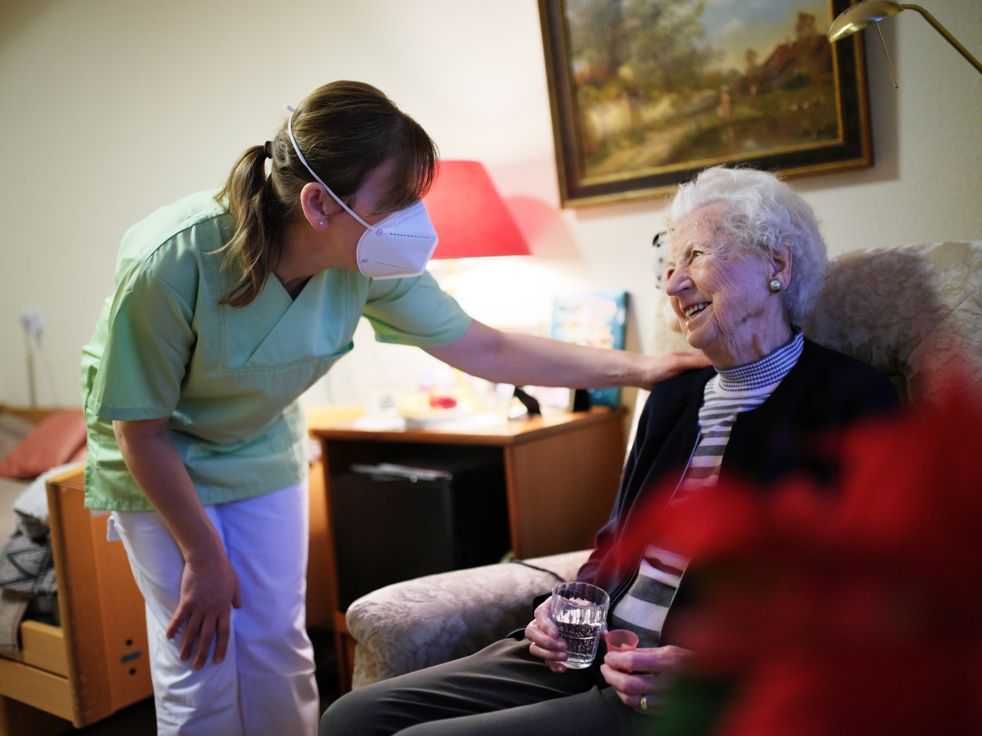 Pflegefachkraft betreut Seniorin in ihrem Zimmer, diese sitzt in einem Sessel beide lächeln sich an
